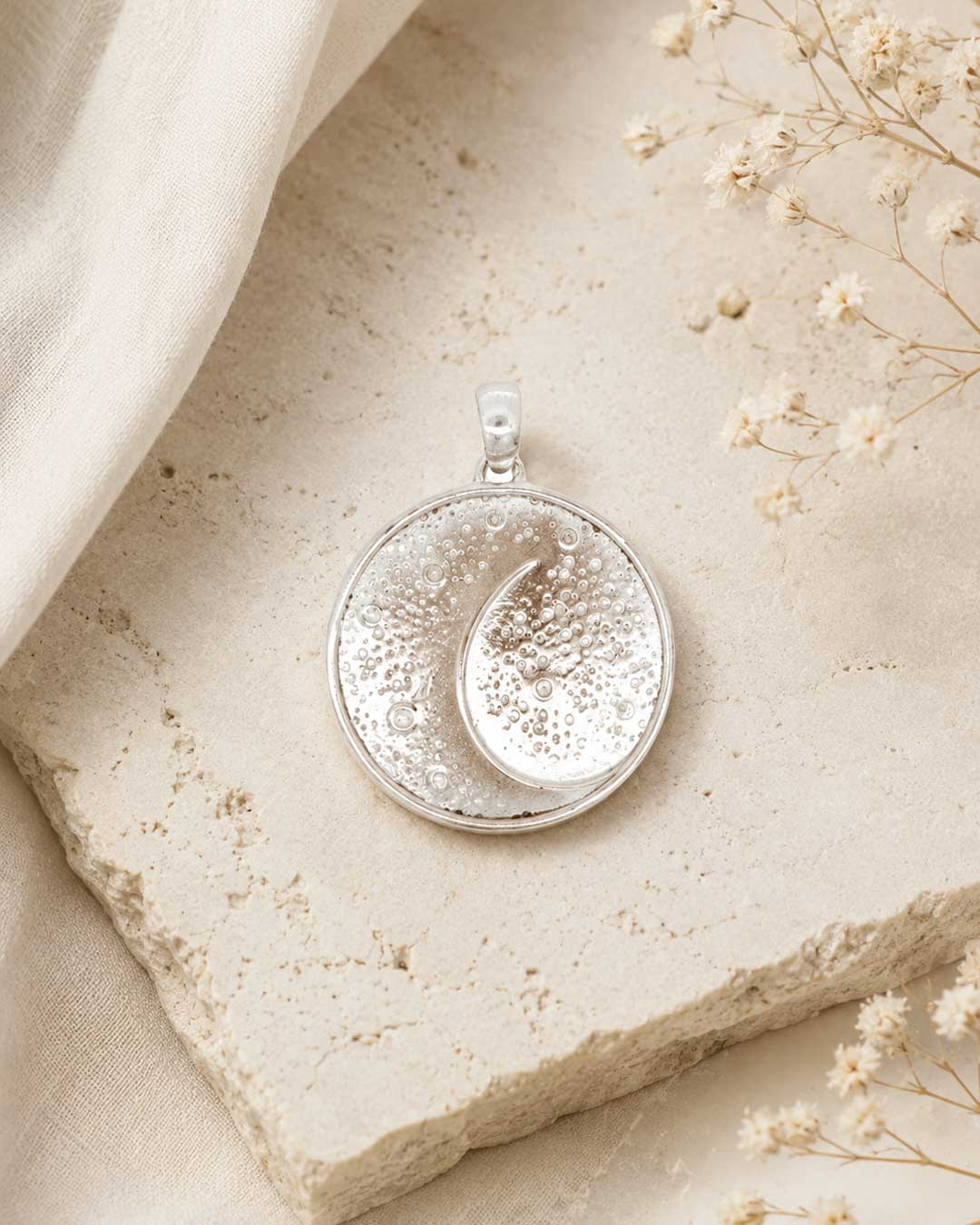 Silver crescent moon pendant laid flat on a stone surface with dried flowers and linen fabric, styled overhead view