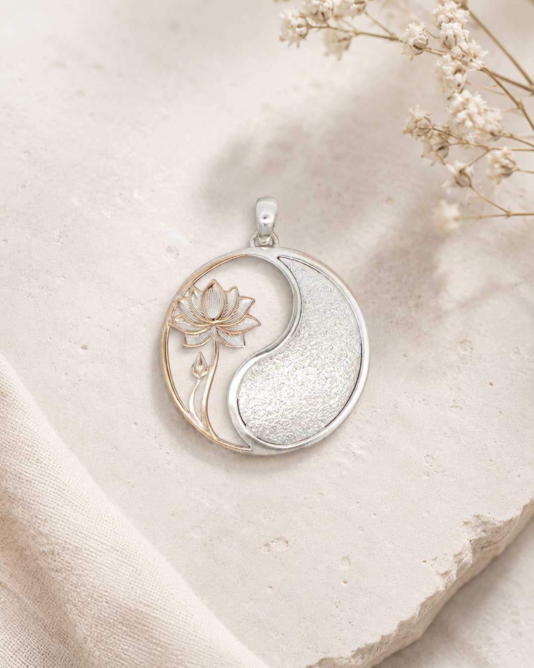 Yin yang lotus pendant resting on rough stone surface with linen fabric and dried baby's breath flowers in the background