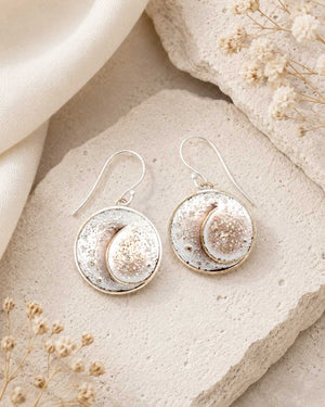 Crescent moon disc earrings laid flat on a stone surface with dried flowers and linen fabric, styled overhead view
