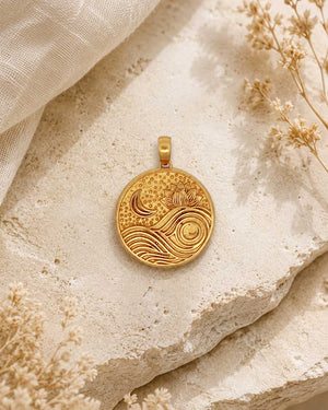 Gold plated brass lotus moon and wave pendant displayed face-up on a textured stone surface with dried botanicals