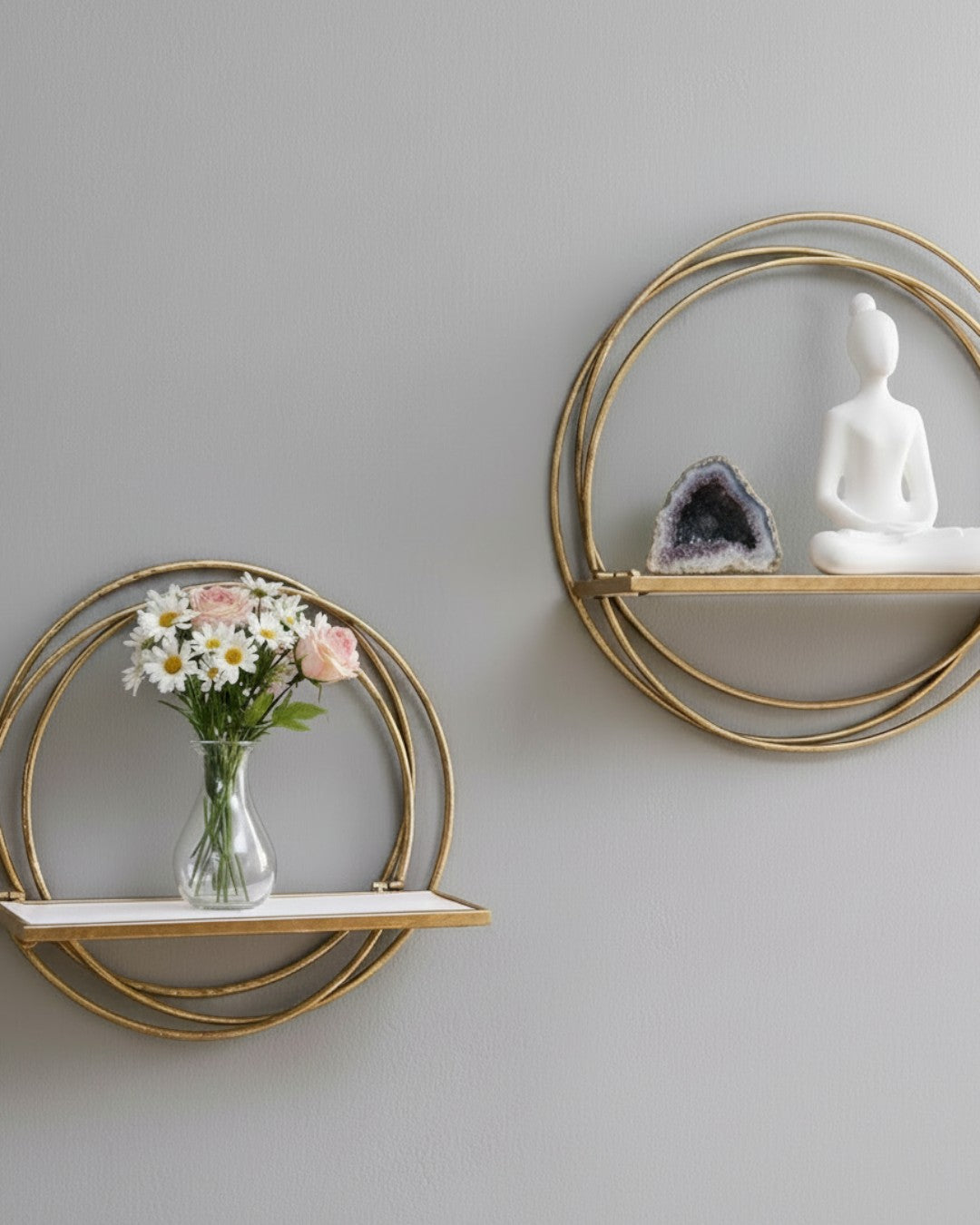 Two golden ring wall shelves on grey wall, lower holding a glass flower vase, upper holding an amethyst geode and white figurine