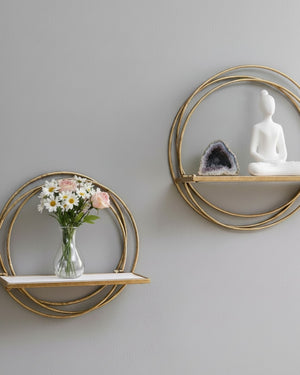 Two golden ring wall shelves on grey wall, lower holding a glass flower vase, upper holding an amethyst geode and white figurine