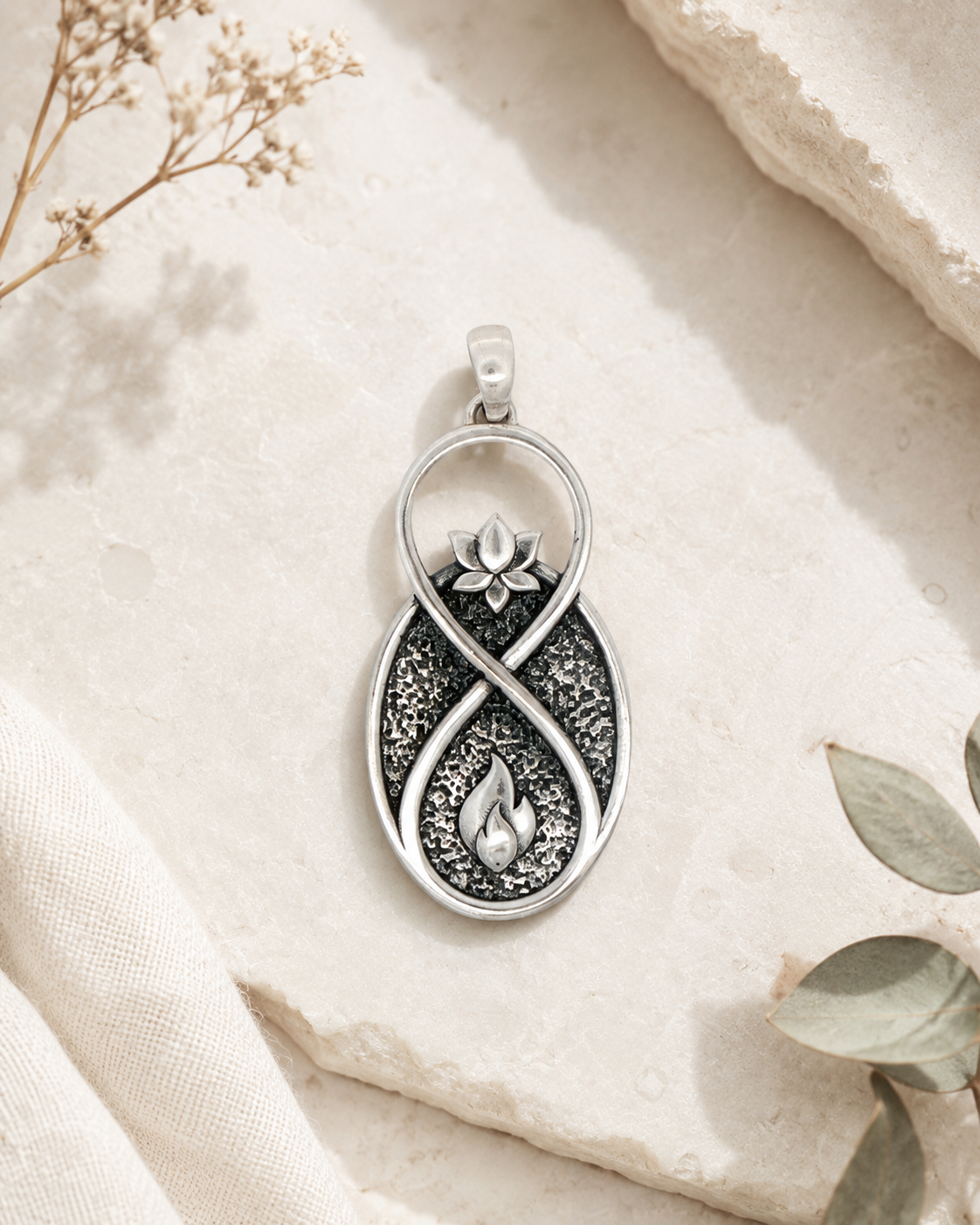 Infinite Lotus Flame Pendant silver charm styled flat on stone surface with dried botanicals and eucalyptus leaves in the background