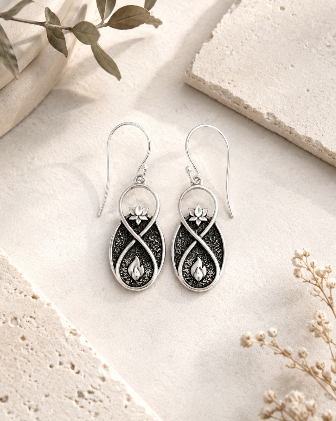 Lotus Infinity Flame earring pair laid flat on stone surface with dried florals and eucalyptus leaves as props