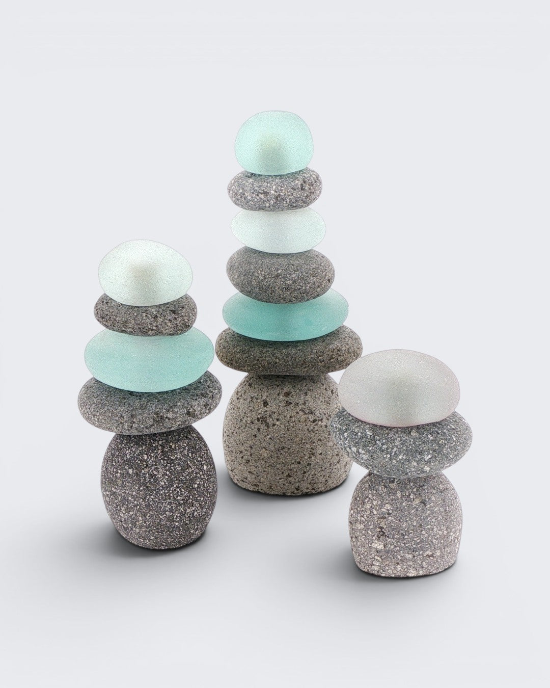 Set of three mini stacked stone cairns with sea glass arranged by height on a light grey background, studio view