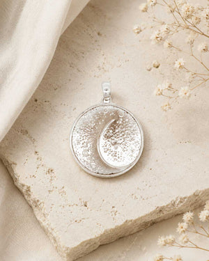 Silver crescent moon pendant laid flat on a stone surface with dried flowers and linen fabric, styled overhead view