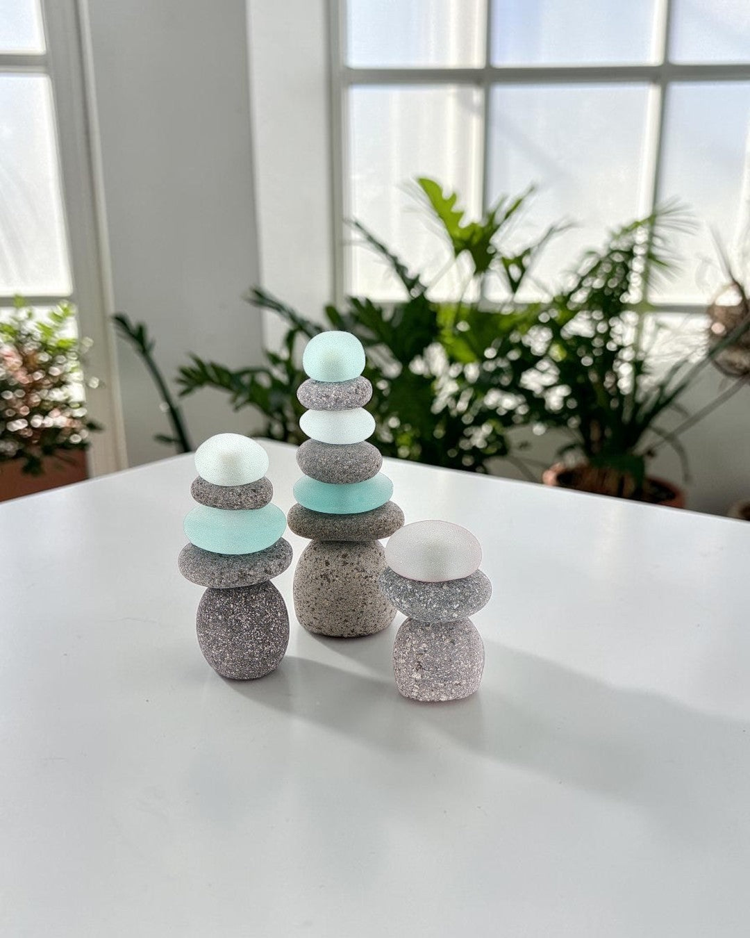Three mini stacked stone cairns with sea glass on a white table, styled with houseplants visible through a window behind